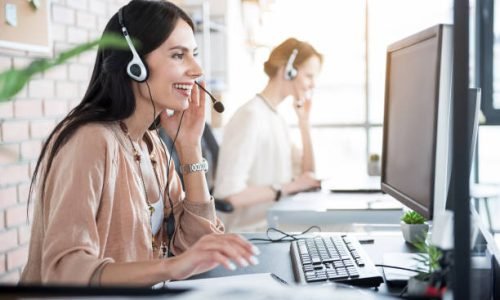 Jolly operator in headphones with microphone is looking at screen of computer and happily smiling. Colleague sitting beside her. Profile